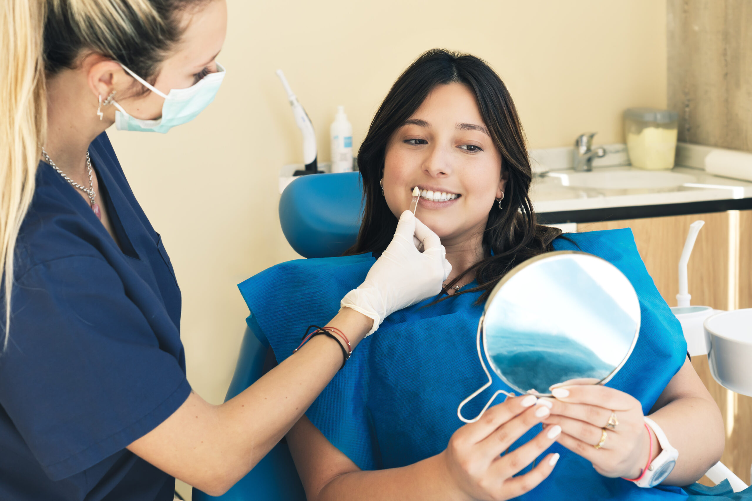 Female dentist checking on female patient teeth color, using tooth enamel scale, dental office interior. The young woman smiles in the dental chair, looking forward to the whitening procedure