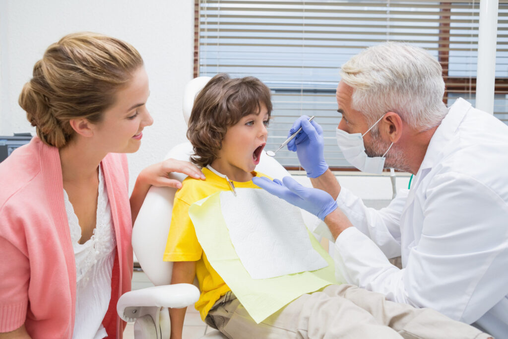 Pediatric dentist examining a little boys teeth with his mother watching at the dental clinic